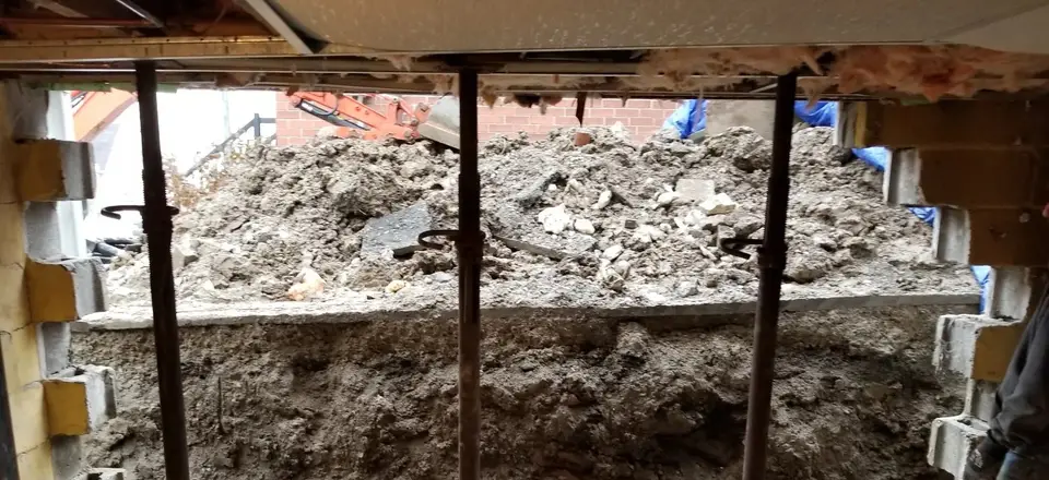 Basement underpinning in progress: soil bench excavated under a block foundation wall with temporary steel shoring